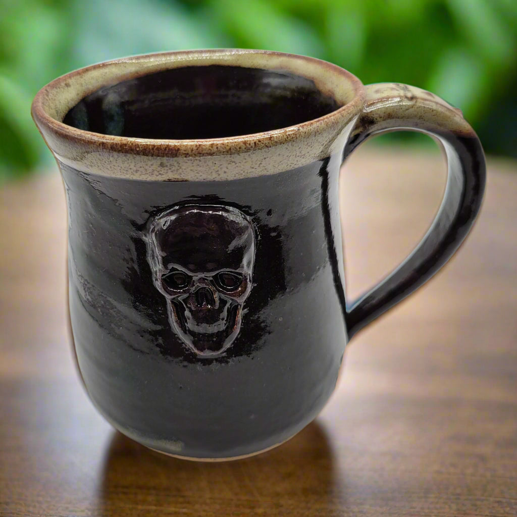 A black ceramic mug with a brown rim and a skull design on the side, handmade in Texas.