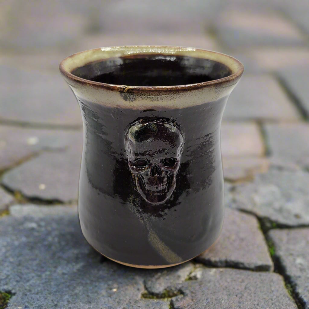 A ceramic mug with a skull design on the front, predominantly dark color with a lighter rim and a skull motif in the center.