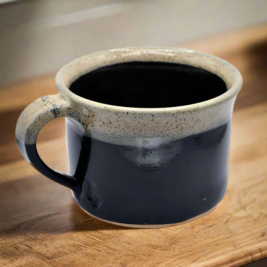 A handmade ceramic mug with a two-tone design, featuring a dark color on the bottom and a speckled light color on the top, with a handle on the left side.
