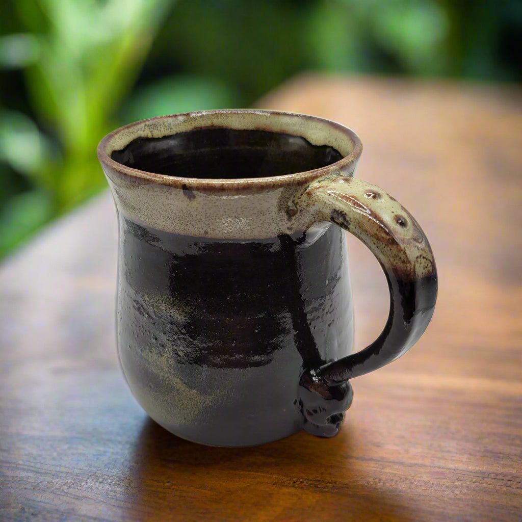 A ceramic mug with a skull design on the handle, predominantly dark color with a lighter rim